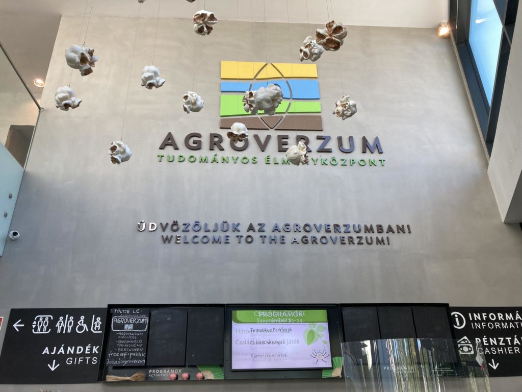 Az Agroverzum Tudományos Múzeum bejárati előcsarnoka. A falon az Agroverzum logó és név magyar és angol nyelven, a mennyezetről pamut növényi dekorációkkal lógnak. Lent található a recepció magyar, angol és német nyelvű ikonokkal és információkkal.