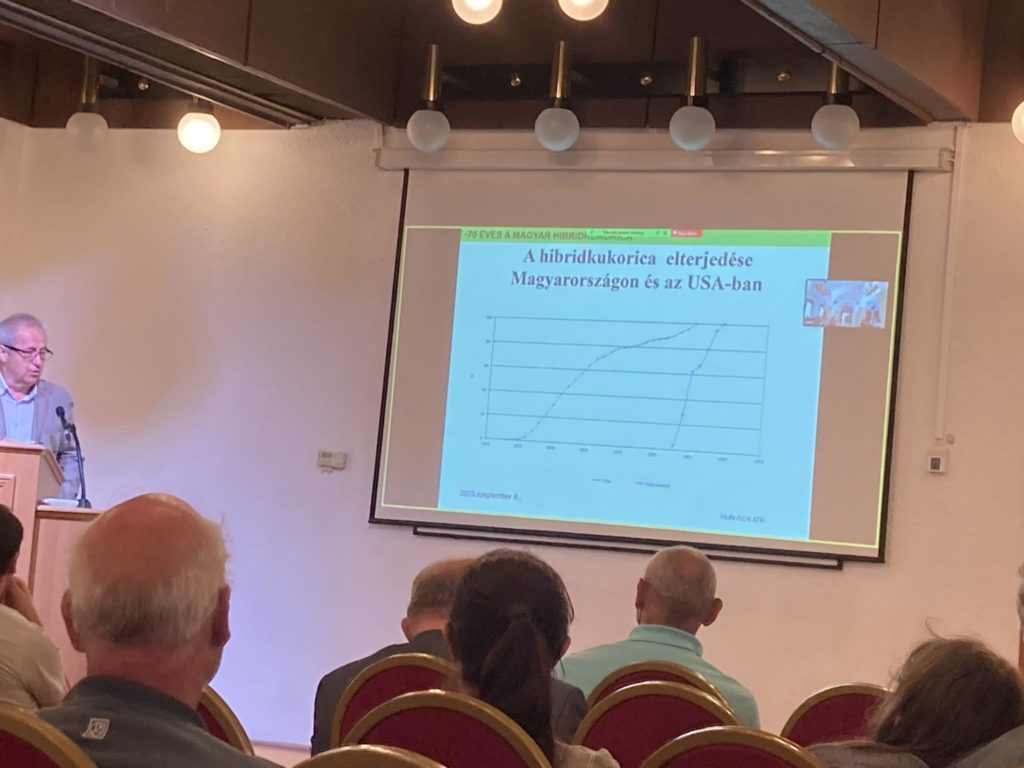 Egy férfi áll az emelvényen, és egy diákat mutat be a hallgatóságnak a konferenciateremben. The slide features a graph titled "A hibridkukorica elterjedese Magyarországon és az USA-ban." A közönség több tagja a képernyő felé néz.