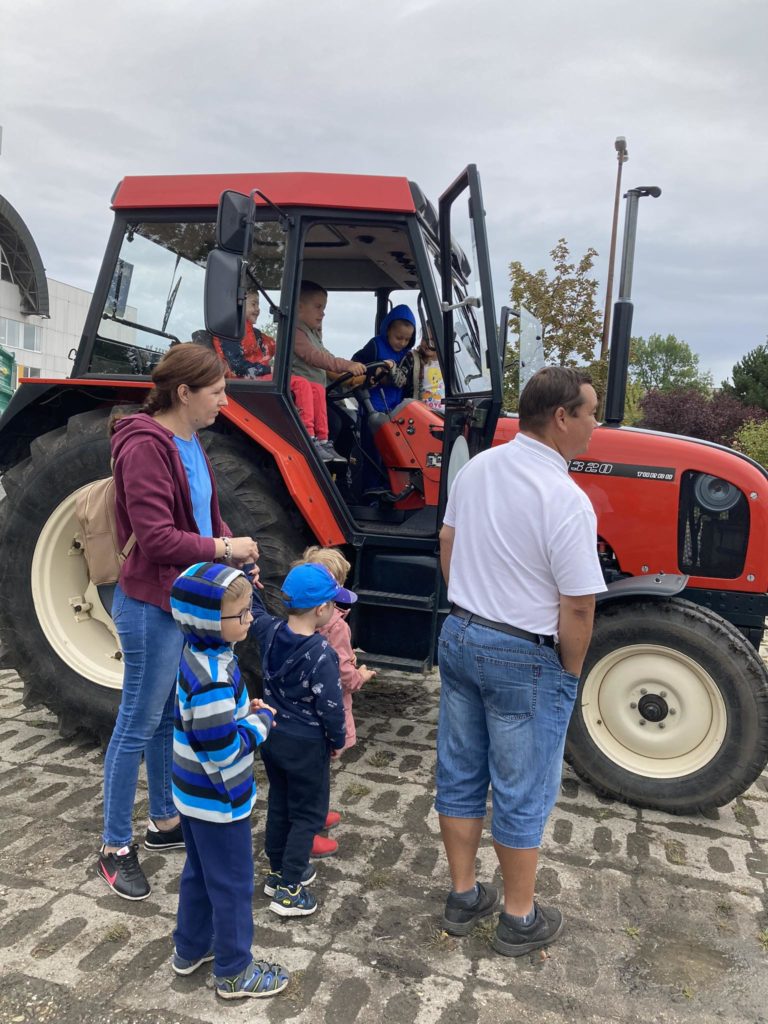 Emberek csoportja, köztük gyerekek és felnőttek, egy piros traktor körül gyűlik össze. A gyerekek színes ruhába vannak öltözve, és néhányan mosolyogva ülnek a traktoron. Úgy tűnik, hogy a jelenet egy szabadtéri, nyilvános területen van, felhős égbolttal.