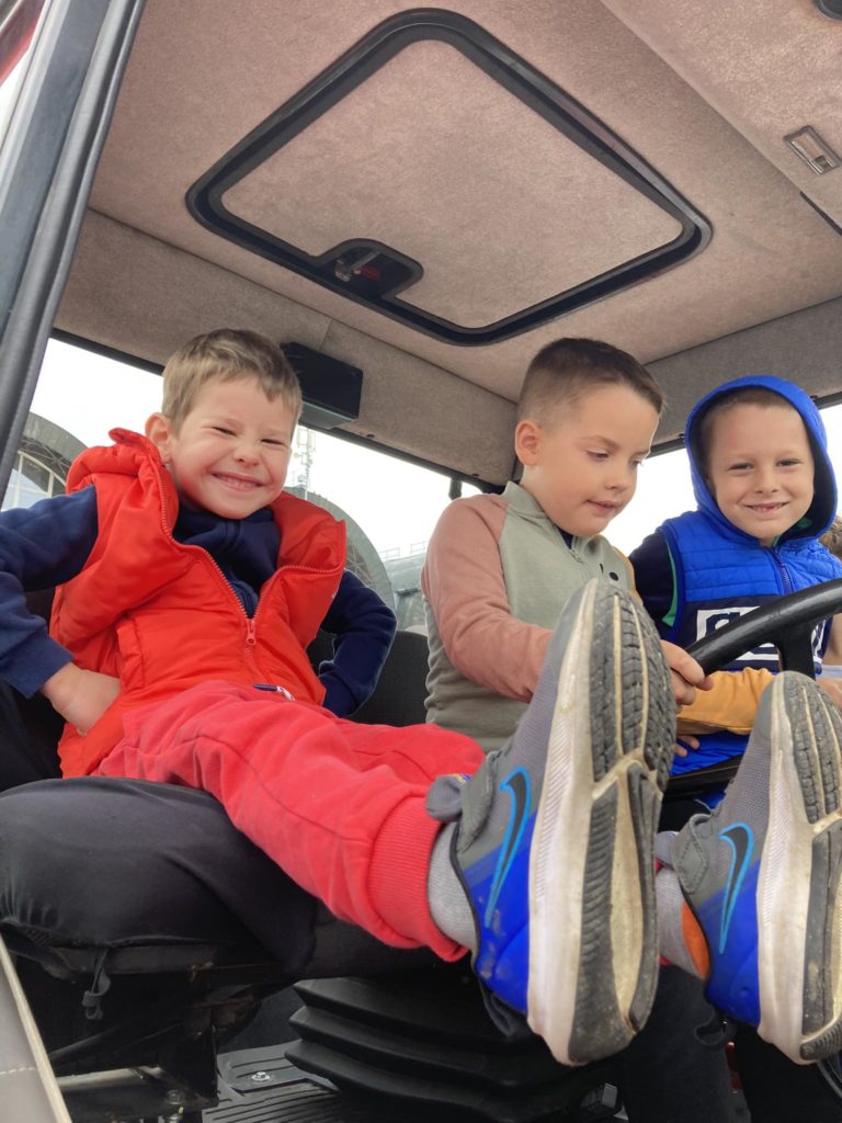 Három fiú ül egy jármű vezetőfülkéjében. A bal oldali fiú vörös ruhába öltözve szélesen vigyorog. A középső, szürke és bézs színű ruhát viselő fiú a kormányra koncentrál. A jobb oldali, kék ruhás fiú mosolyogva nézi a többieket.