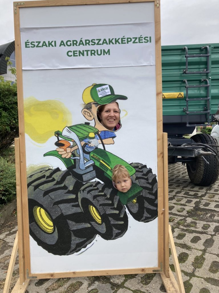 Egy nő és egy gyerek pózol egy kivágott tábla mögött, lyukak egy zöld traktort vezető karikatúra farmerhez igazodva. A tábla „Északi Agrárszakképzési Centrum” felirattal van ellátva. A szabadban vannak, egy zöld pótkocsi mellett.