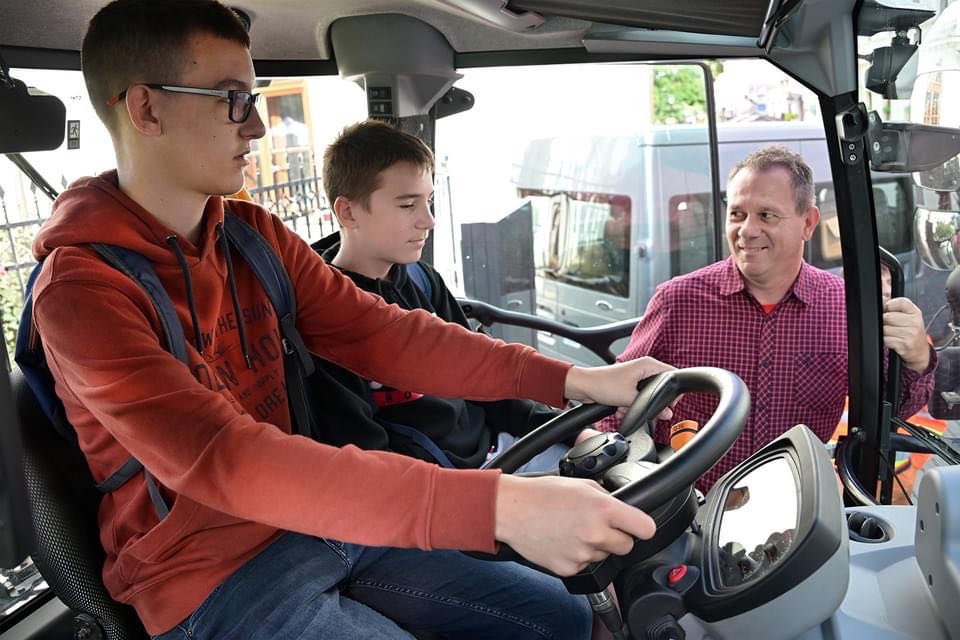 Két fiatal fiú ül egy jármű első ülésén, egyikük a kormányt fogja. Egy idősebb férfi mosolyogva áll a nyitott vezetőajtó előtt, és beszélget velük. A jármű belseje és az ablakok tükröződései láthatók.