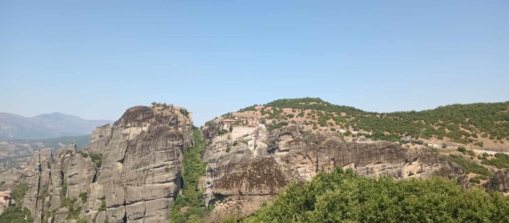 A képen a görögországi Meteora panorámája látható, hatalmas sziklaoszlopok tetején álló kolostorokkal. A sziklás tájat zöld növényzet veszi körül a tiszta kék ég alatt. A jelenet kiemeli a lenyűgöző természeti képződményeket és az ősi építészetet.