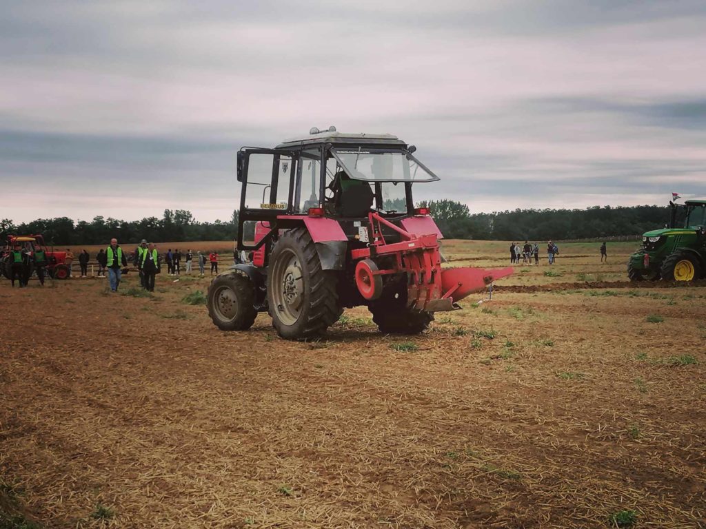 Egy piros traktor parkolt egy mezőn, több emberrel és egy másik traktorral a háttérben. Az ég borús, és a mező száraznak tűnik fűfoltokkal. A háttérben lévő személyek megfigyelik vagy dolgoznak.