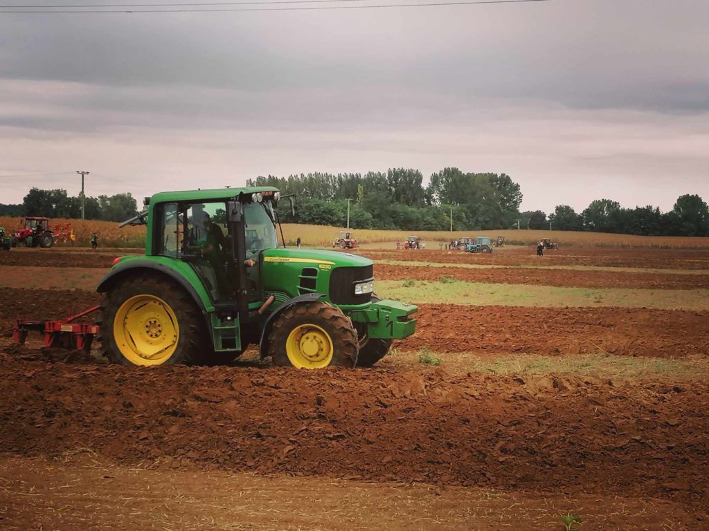 Egy zöld, sárga kerekű traktor szánt egy hatalmas, barna mezőt a borús égbolt alatt. A távolban más gépek és járművek láthatók. A mezőt zöld fák veszik körül.