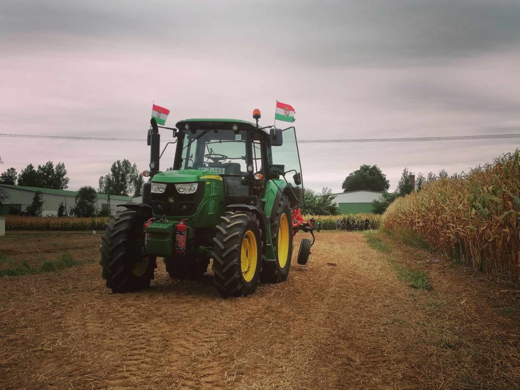 Egy zöld traktor, rajta zászlókkal, egy földúton parkol a kukoricatáblán, felhős ég alatt. A környező táj több kukoricatáblát és néhány távoli fát mutat. A traktor jól láthatóan a kép közepén helyezkedik el.