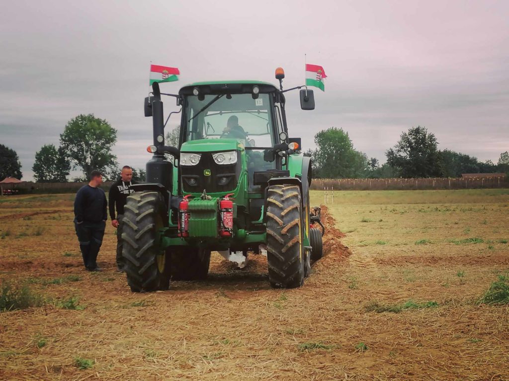 Két ember áll egy zöld traktor mellett egy mezőn. A nagy abroncsokkal és magyar zászlókkal felszerelt traktor átvág a tarlóval borított talajon. Az ég borús, a háttérben fák sorakoznak.