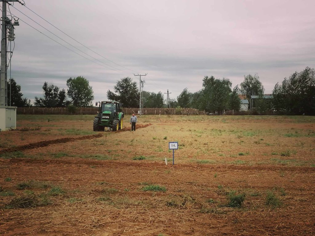 Egy traktor műveli a talajt egy nagy, gyéren növényzett mezőn a felhős égbolt alatt. Két ember sétál mögötte. Az előtérben egy kis számozott tábla, „14” van elhelyezve. A háttérben fák és villanyvezetékek láthatók.