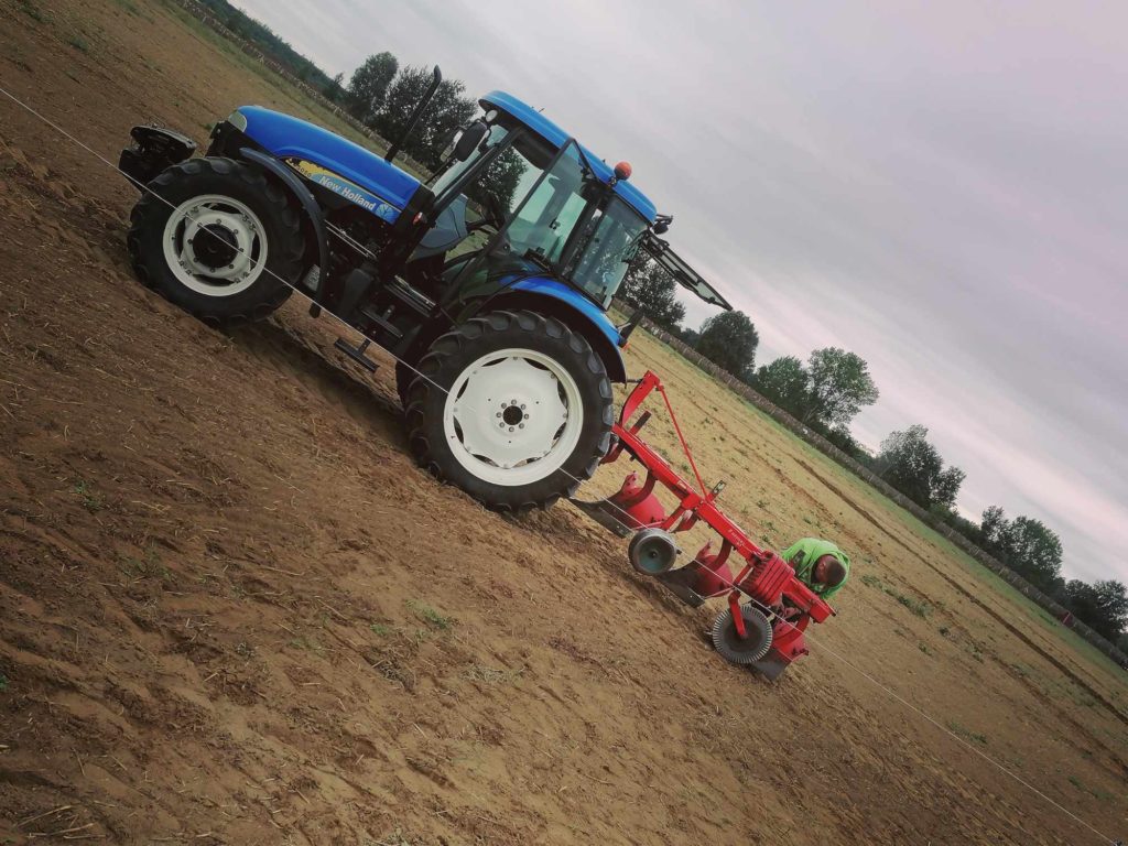 Egy kék traktor hatalmas, barna mezőt szánt a borús égbolt alatt. A traktor piros mezőgazdasági gépet húz, amelyet a talaj megmunkálására terveztek. A háttérben fák és néhány távoli épület látható.