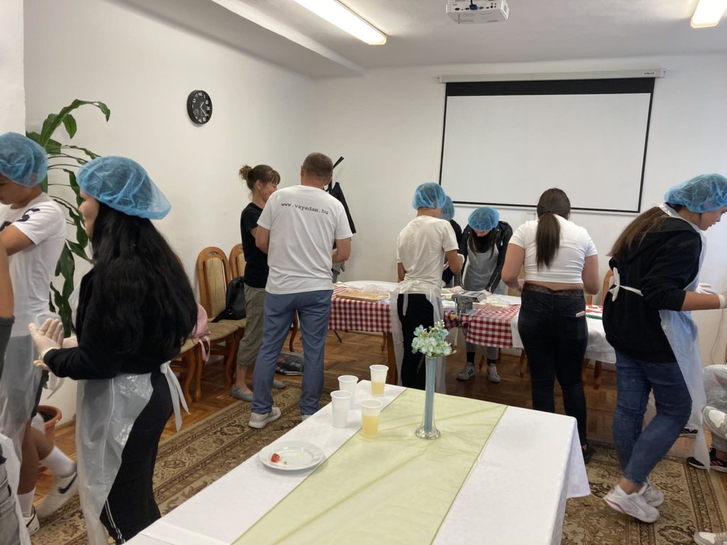 Kék hajhálót és fehér kesztyűt viselő embercsoport asztalok körül áll egy fehér falú és órával rendelkező szobában. Úgy tűnik, hogy ételkészítési tevékenységet folytatnak, az összetevőket és az edényeket az asztalokra rakták.
