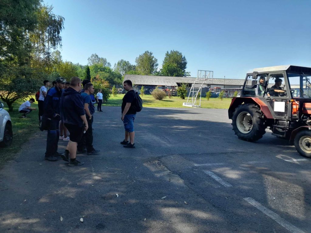 Emberek egy csoportja, főként férfiak, egy parkolóban áll egy fehér autó és néhány fa mellett. Jobb oldalról traktor közeledik. A háttérben több ember, zöld növényzet és egy alacsony épület látható a tiszta kék ég alatt.