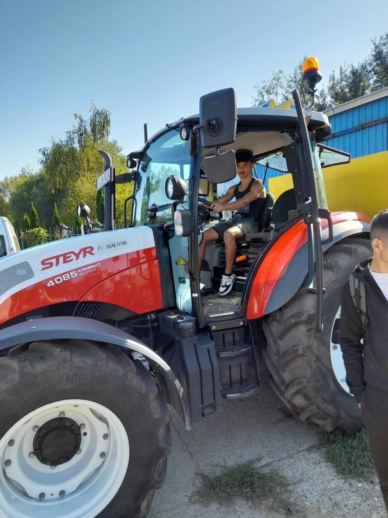 Egy személy ül egy piros-fehér Steyr 4085 Kompakt traktorban, egyik kezével a kormányon. A traktor a szabadban parkol egy napsütéses napon. Egy másik személy áll a közelben, részben látható a kép jobb szélén. A háttérben fák és egy kék épület látható.