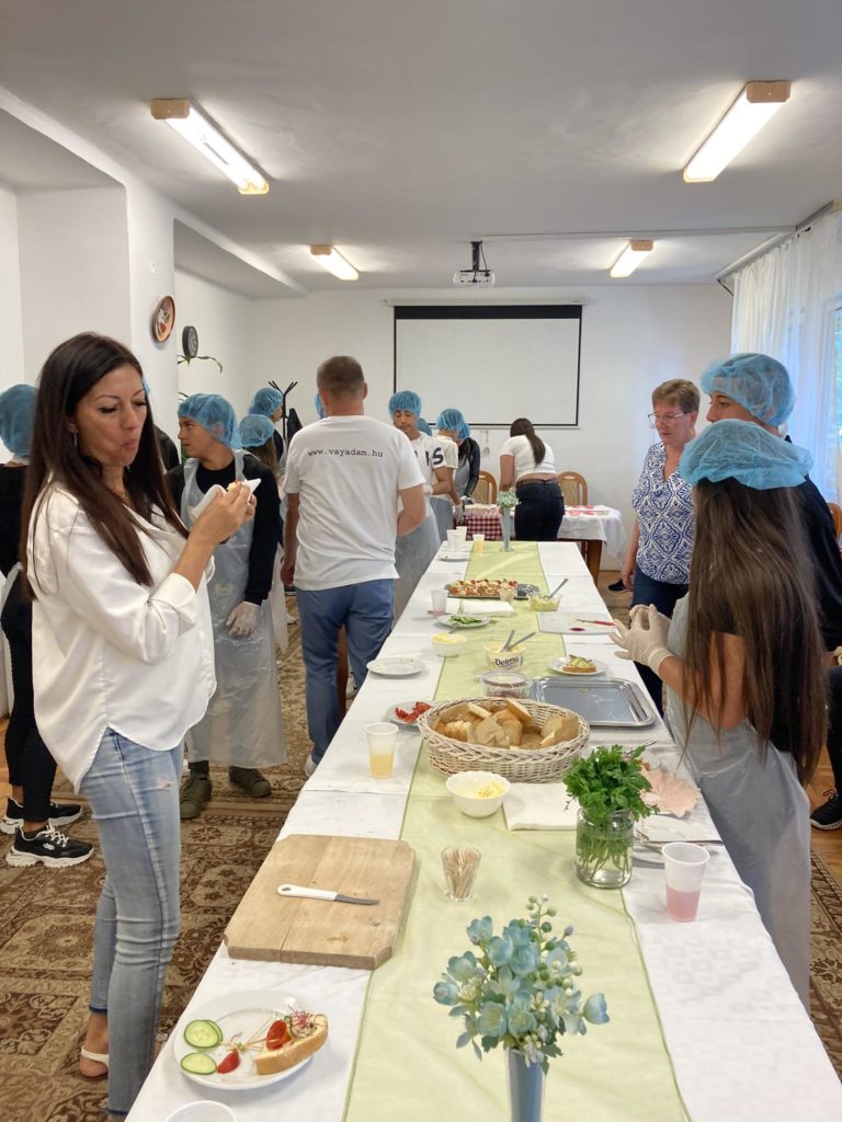 Egy világos szobában egy csoport ember gyűlt össze egy asztal körül, ahol különféle ételeket adtak. Néhányan kék hajhálót és kötényt viselnek, ami főzési tevékenységre utal. Egy nő az előtérben eszik. Az asztalon növények és dísztárgyak vannak.
