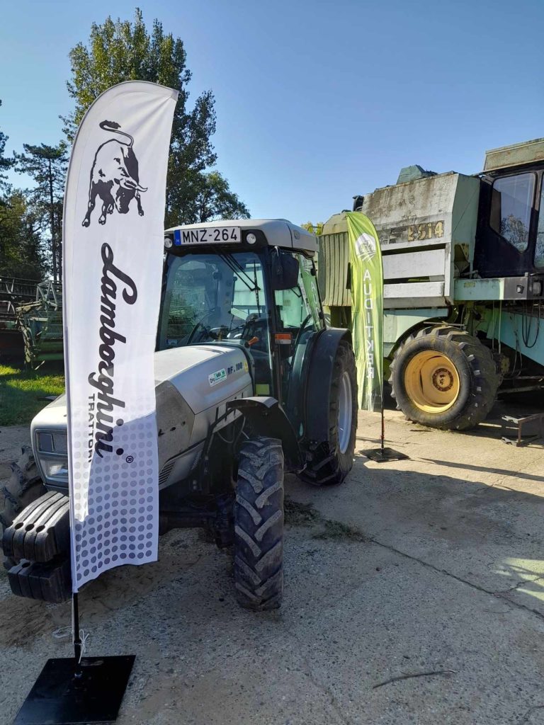 Egy fehér Lamborghini márkájú traktor parkolt betonfelületen zöld-fehér transzparensek mellett. A háttérben egy nagy, régebbi mezőgazdasági gép látható. Tiszta az ég, a háttérben fák állnak.