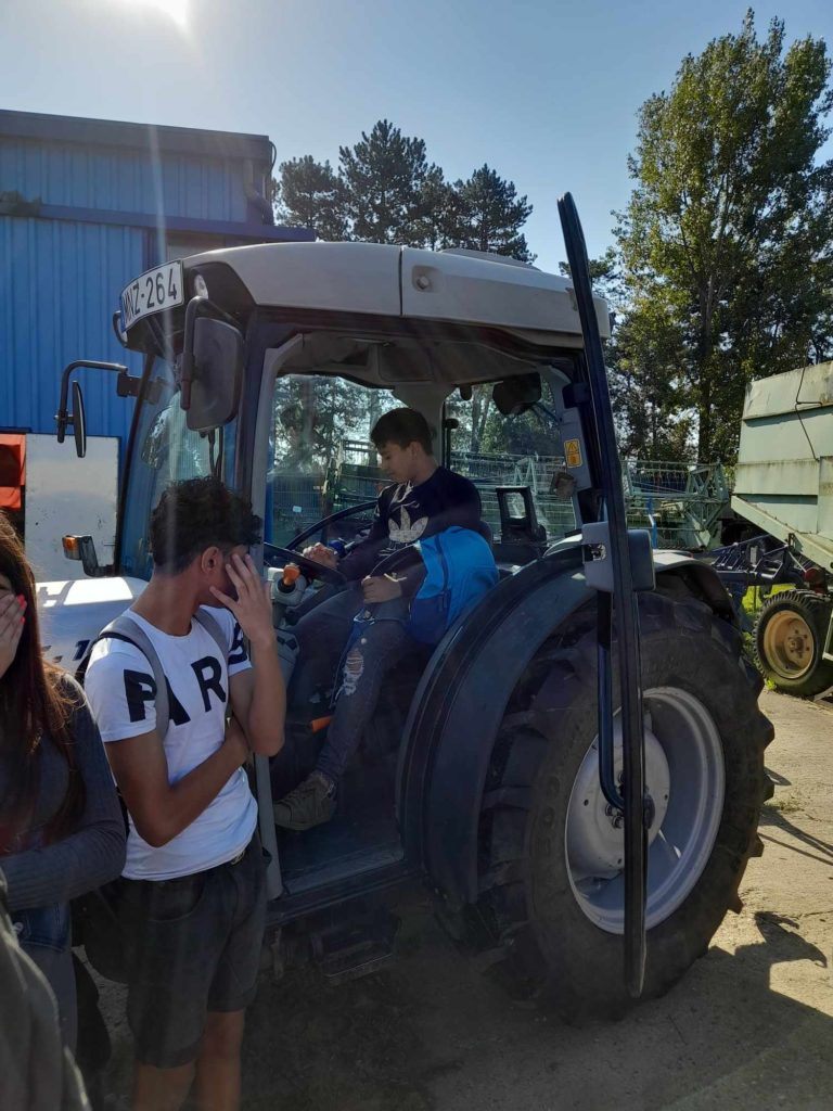 Egy személy ül egy parkoló traktor vezetőfülkéjében, kezével a kormányon, és esetleg vezetést szimulál. Három másik személy a traktoron kívül áll, ketten a kezükkel eltakarják az arcukat. A háttérben fák és épületek láthatók.