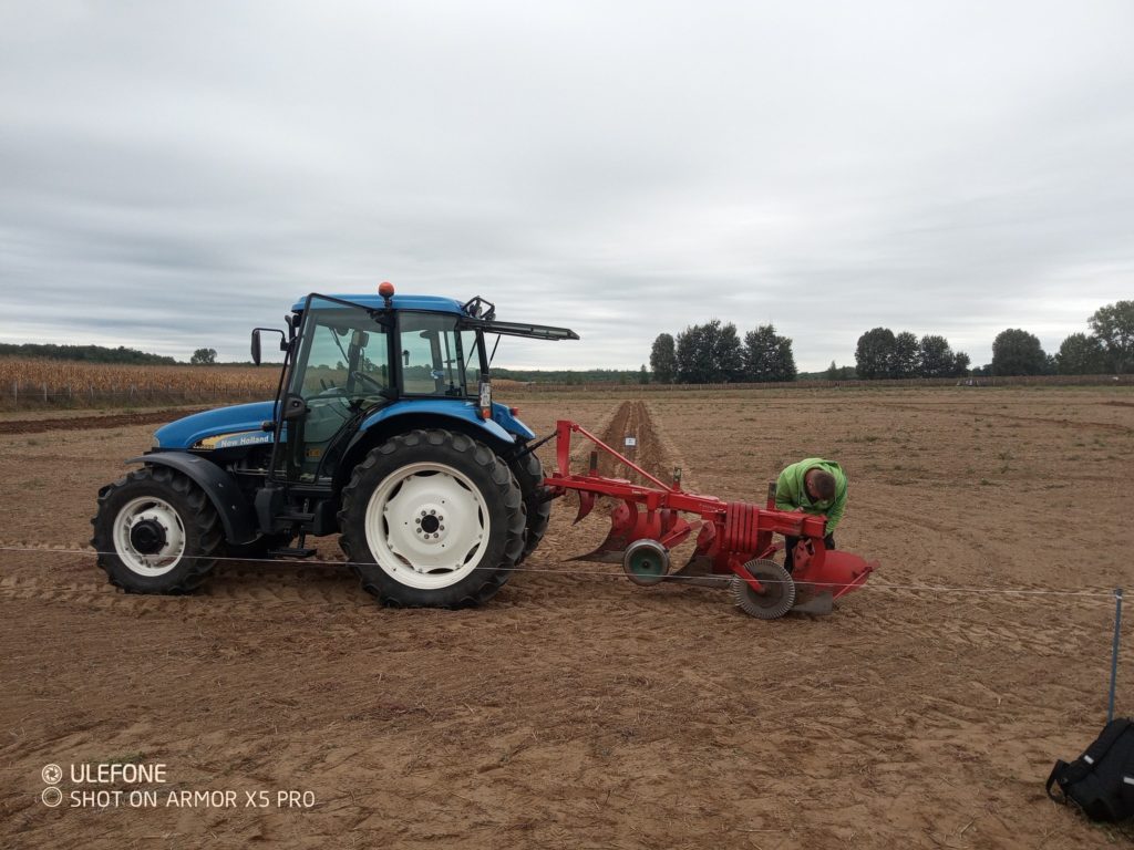 Egy traktor egy piros mezőgazdasági gépet húz egy felszántott területen a felhős ég alatt. Egy személy a gép közelében áll, és esetleg beállítja vagy felügyeli. A háttérben fák és egy szántóföld látható. A sarokban lévő szöveg azt jelzi, hogy a fénykép Ulefone Armor X5 Pro-val készült.