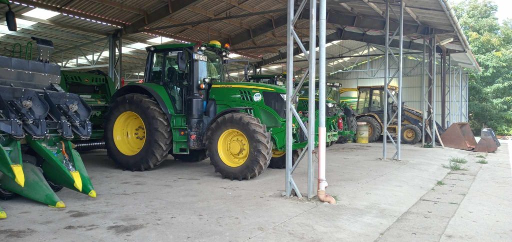 Zöld és sárga traktorok sora parkol egy nagy fémtetős fészerben. A fészer nyitott előlappal rendelkezik, és fémgerendák támasztják alá. A traktorok különböző mezőgazdasági berendezések mellett helyezkednek el. A háttérben fák láthatók.