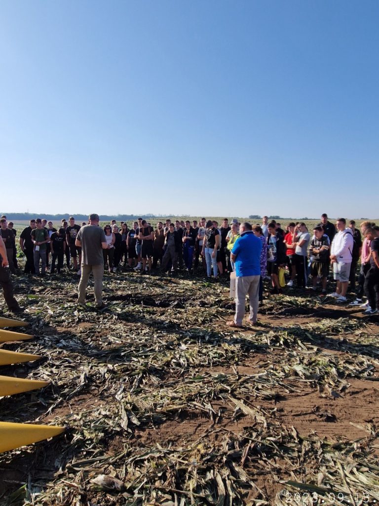 Egy csoport ember áll együtt egy mezőn a tiszta kék ég alatt. Úgy tűnik, hogy egy előttük beszélő személyt hallgatnak. A talajt száraz kukoricaszár borítja, ami a közelmúltbeli betakarításra utal. Néhány nagy sárga mezőgazdasági gép részben látható.