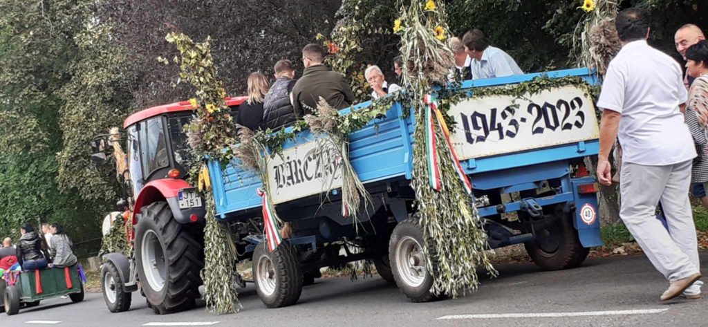 Egy piros traktor lombokkal és virágokkal díszített kék pótkocsit húz egy megemlékező felvonuláson. Az előzetesen „1943-2023” felirat látható. Emberek utaznak az utánfutóban, nézők pedig az út mentén. A háttérben fák sorakoznak.