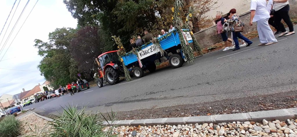 Utcai felvonulás, amelyen egy traktor húz egy kék pótkocsit, amely díszekkel és egy "La Raclette" táblával díszített, majd egy csoport ember sétál. A bámészkodók az út mentén állnak, a háttérben több fa és épület is látható.