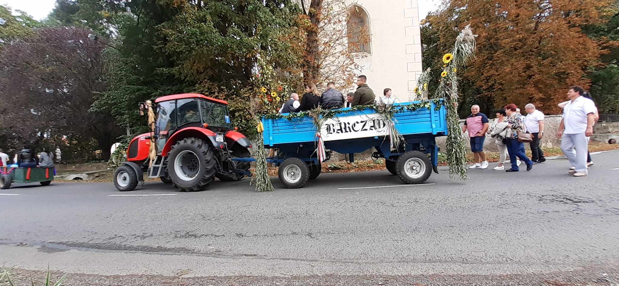 Egy piros traktor húz egy kék, virágokkal és zölddel díszített pótkocsit, amelyen több ember ül és áll. Az előzetesen lévő transzparens „Ranica” felirattal. A bámészkodók a közelben, az utcán állnak, fákkal és egy épülettel a háttérben.