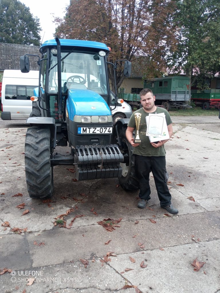 Egy férfi áll egy kék traktor mellett, két trófeával és egy bizonyítvánnyal. Zöld inget és sötét nadrágot visel. Fák és egy fehér furgon a háttérben. Úgy tűnik, vidéki vagy tanyasi környezet.