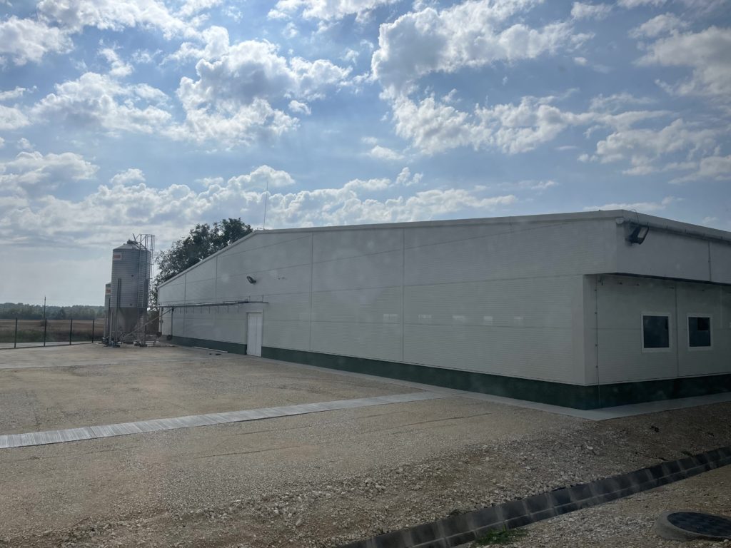 Egy nagy, fehér, földszintes ipari épület áll kavicsos telken, részben felhős égbolt alatt. Az épület alján zöld burkolat található. A háttérben silók és a horizontig nyúló nyílt mezők láthatók.