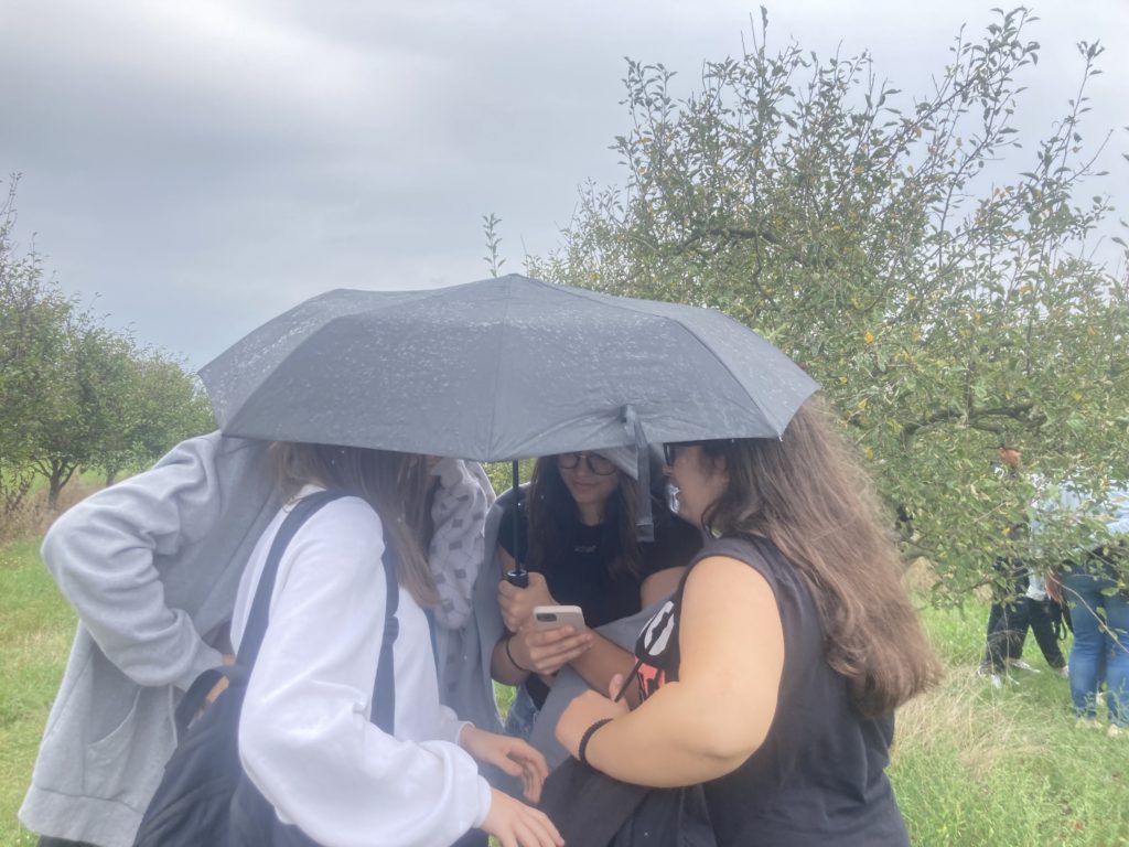 Négy ember húzódik meg egyetlen fekete esernyő alatt egy esős gyümölcsösben. Szorosan összegyűltek, és az egyiküknél tartott telefont nézik. A háttérben zöld fű, fák és szürke, borult égbolt látható.
