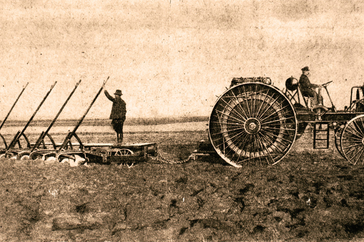 Vintage fénykép a 20. század eleji gazdálkodásról. Egy nagy, küllős kerekekkel rendelkező traktor többkéses ekét húz a nyílt területen. Két gazda dolgozik: az egyik a traktort vezeti, míg a másik az ekén áll és vezeti azt. A jelenet szépia tónusú.
