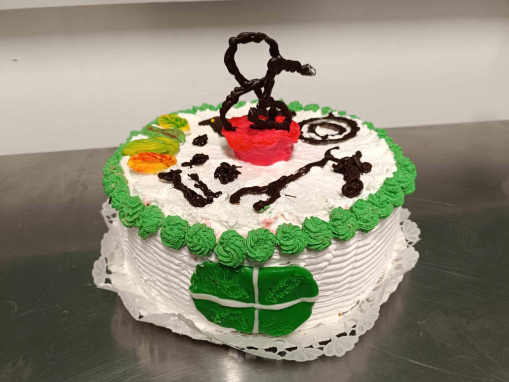 Kerek, fehér torta, szélein zöld cukormázzal, tetején válogatott színes díszítéssel. A közepén egy vörös és fekete absztrakt szobor látható. A torta ezüst felületen van kihelyezve, alatta csipke alátéttel.