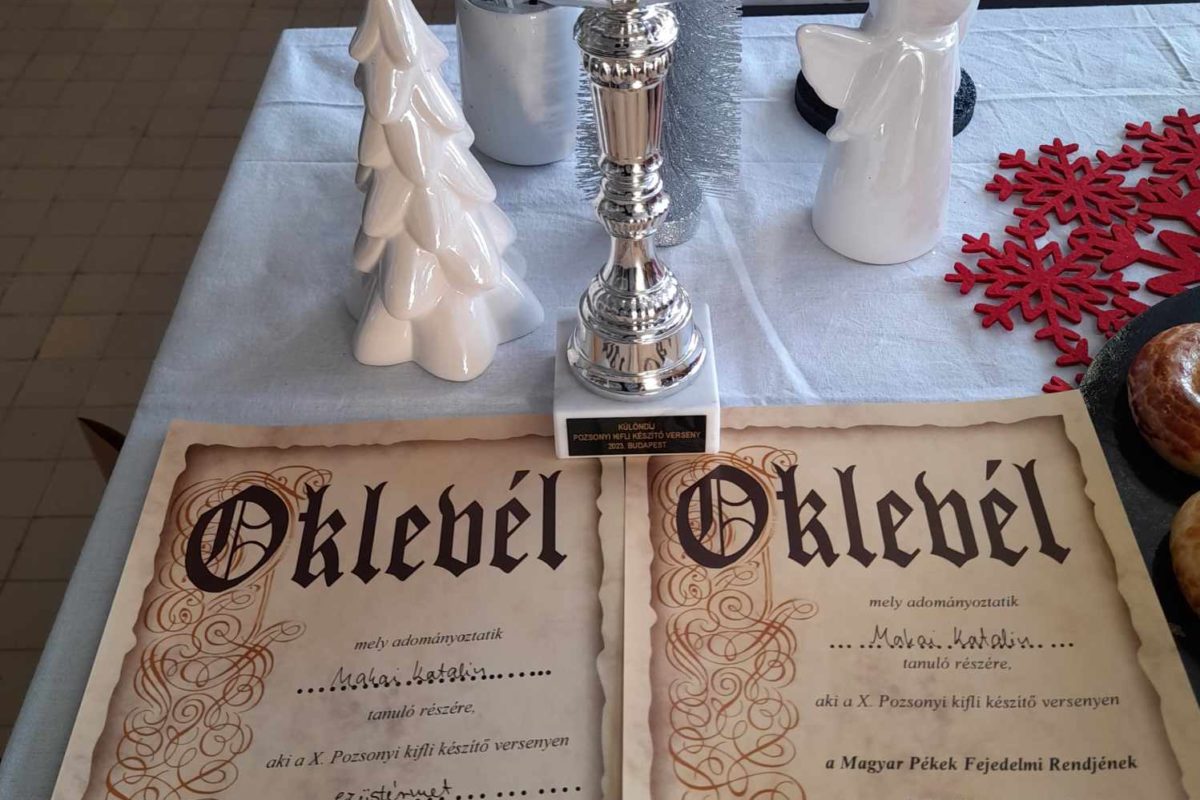 Egy asztalon egy ezüst trófea és két magyar „Oklevel” oklevél látható. A trófea mellett egy fehér kerámia karácsonyfa és egy figura. Az asztal fehér terítővel van lefedve, amin egy kockás piros kendő részben látható.