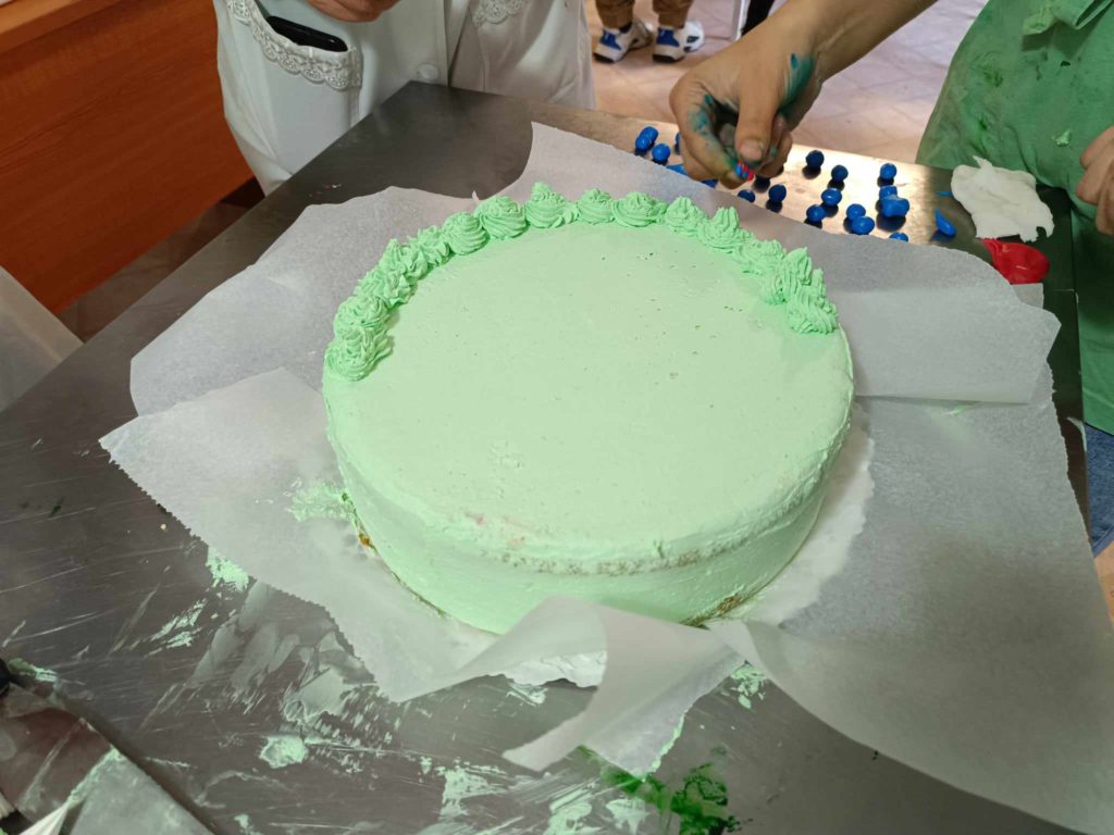 Egy személy világoszöld cukormázas tortát díszít, szélén zöld cukormáz örvényl. A háttérben kék-fehér fondant díszítések láthatók. A jelenet egy konyhában vagy pékségben játszódik.