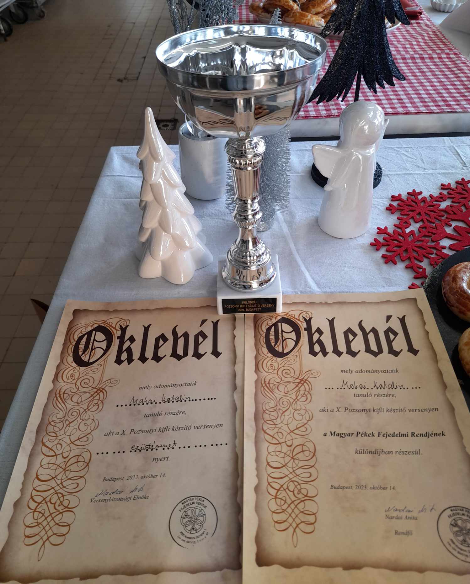 Egy asztalon egy ezüst trófea és két magyar „Oklevel” oklevél látható. A trófea mellett egy fehér kerámia karácsonyfa és egy figura. Az asztal fehér terítővel van lefedve, amin egy kockás piros kendő részben látható.