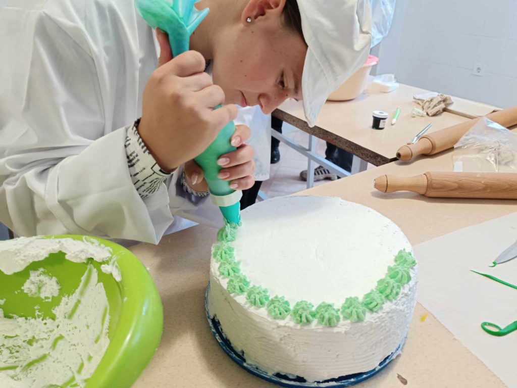 Egy fehér kalapot és kabátot viselő személy egy fehér tortát zöld cukormázzal díszít egy zsák segítségével. A torta egy asztalon van, a közelben különféle sütőeszközökkel, köztük sodrófával, cukormázzal ellátott tálkával és tortadíszítő eszközökkel. Úgy tűnik, a személy a feladatára összpontosít.