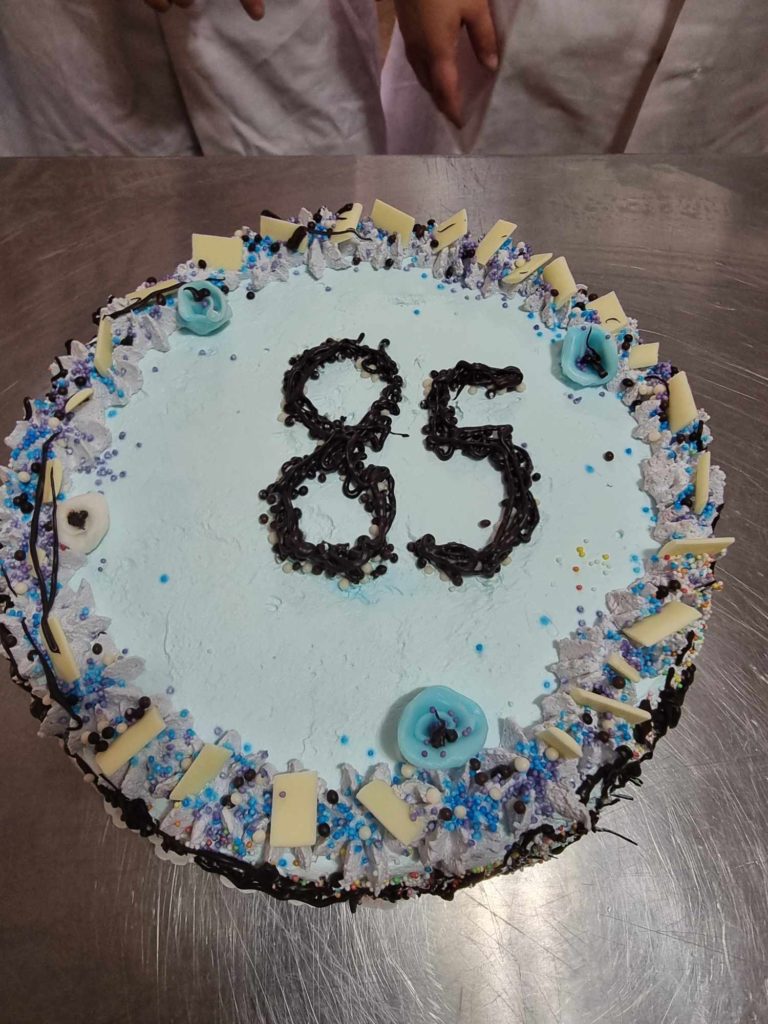 Egy világoskék cukormázas kerek torta tetejére étcsokoládéval a „85” szám van írva. A tortát fehér csokoládédarabkák díszítik, a szélein kék és ezüst szórással. A torta mögött a háttérben két kéz látható.