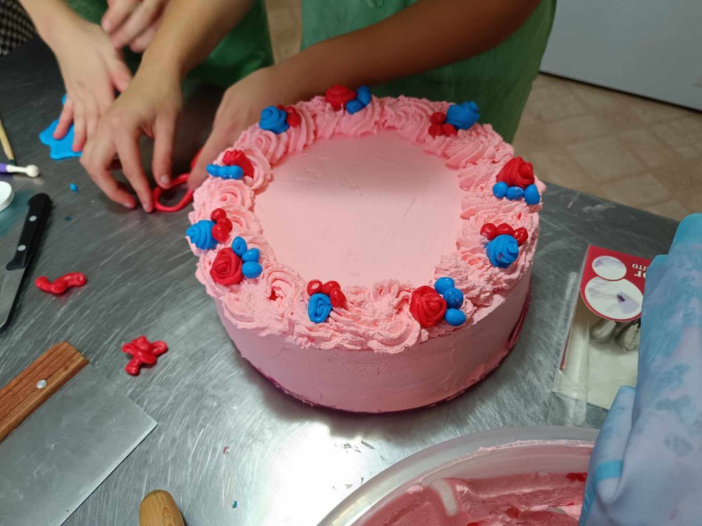 Egy személy egy kerek tortát sima rózsaszín cukormázzal díszít. A tortát a szélein rózsaszín és kék habvirágok díszítik. A közelben egy másik ember keze vörös fondanttal dolgozik egy rozsdamentes acél felületen. Különböző eszközök és dekorációk vannak szétszórva.