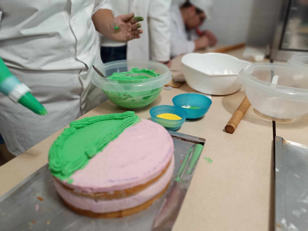 Egy fehér köpenyt viselő személy egy tortát díszít zöld cukormázzal egy zsák segítségével. A torta egy tálcán fekszik, részben rózsaszín cukormázzal borítva. Az asztalon több cukormázt és egyéb hozzávalókat tartalmazó tál, sodrófával együtt.