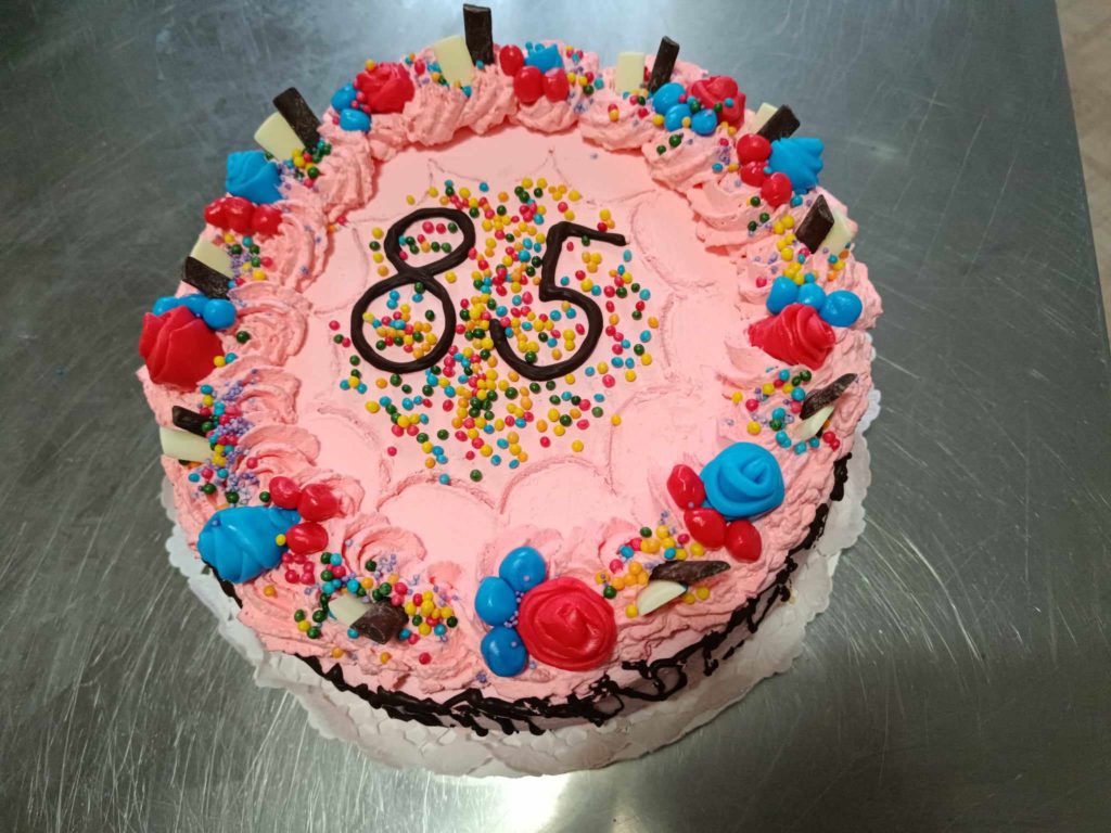 Rózsaszín cukormázzal díszített kerek torta, színes szórással, rozettákkal és cukorkadarabokkal díszítve. A torta tetején két csokoládészám, „85” található, jelezve a 85. születésnap ünneplését. A torta ezüst felületen ül.