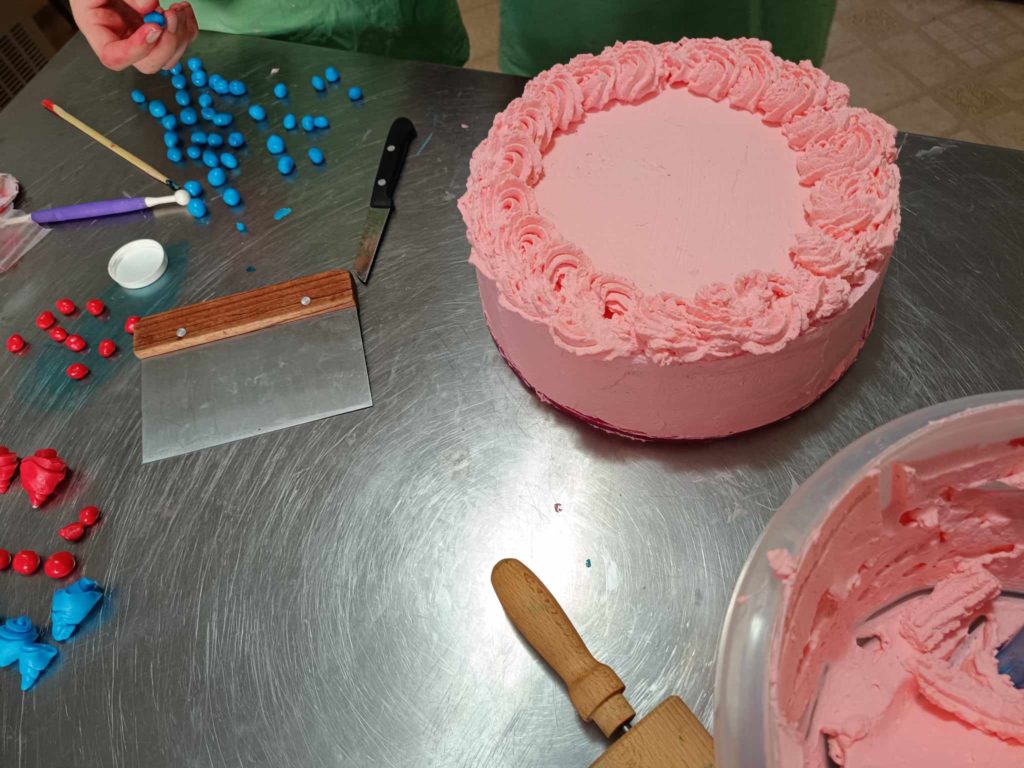 Fém felületen egy részben díszített kerek rózsaszín torta ül. A torta körül különböző díszítőeszközök, spatula, valamint piros és kék gömb alakú dekorációk találhatók. A törzstől lefelé látható személy bottal dekorációt rak össze.