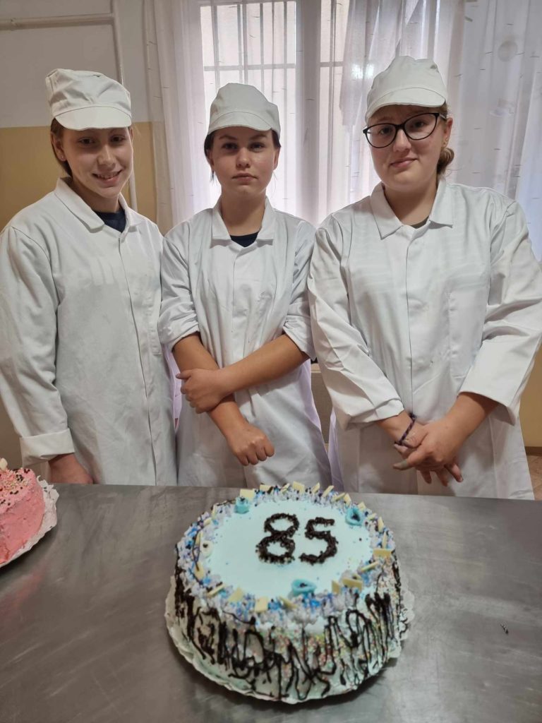 Három fehér egyenruhába és sapkába öltözött személy áll egy fémfelületen díszített torta mögött. A tortán fekete cukormázzal a „85” szám szerepel. A háttérben fehér függönyök és sárga fal látható.