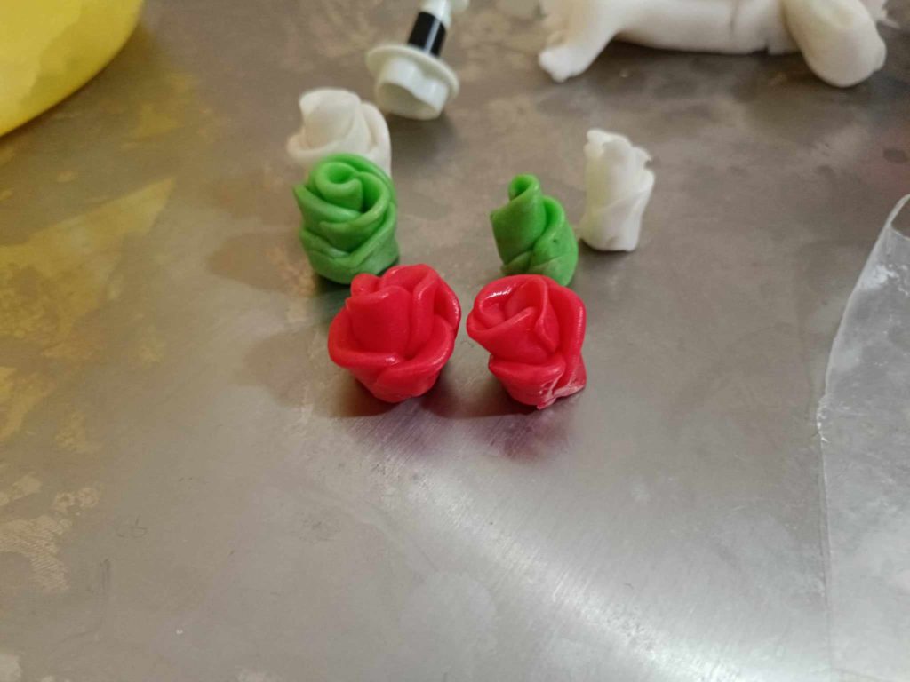 Közeli kép rózsa alakú fondant tortadíszekről. Két vörös rózsa látható az előtérben, két zöld rózsa és két fehér rózsa enyhén elmosódott a háttérben. Egy kis fondant eszköz és néhány fondant darab is látható.