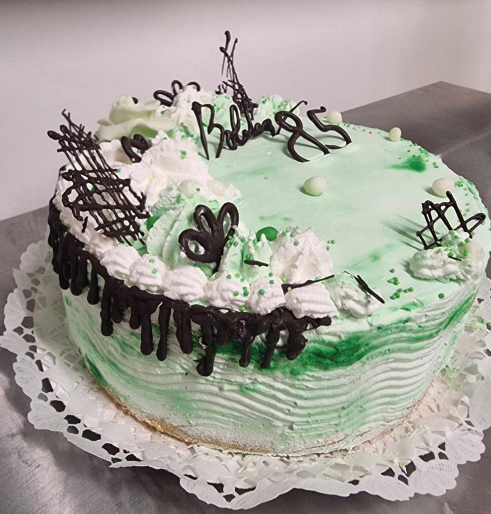 Egy gyönyörűen díszített zöld-fehér születésnapi torta ül az asztalon. A tortán tejszínhab, csokoládé díszítés és a tetején a "85" szám látható. A tortát kerek csipkemintás papírszalvétára tesszük.