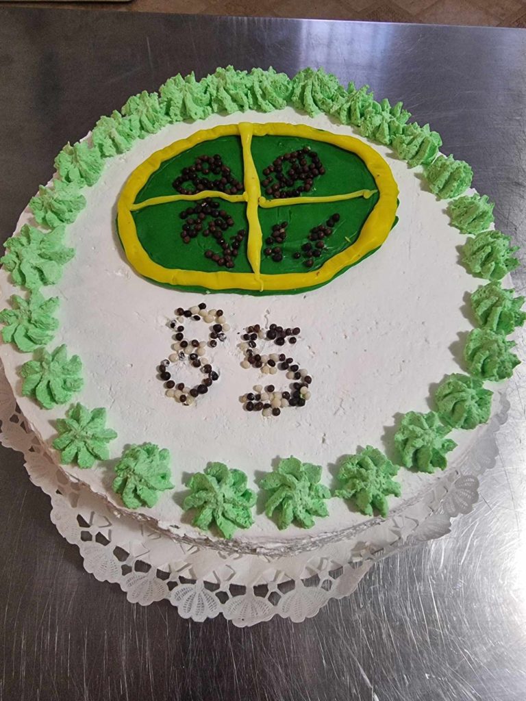 A díszített torta szélén zöld cukormáz látható, középen kis sötét gyöngyökkel díszített sárga és zöld mintával. A „35” szám középre van írva hasonló gyöngyökkel. A torta csipkemintás papírszalvétán és fémes felületen nyugszik.