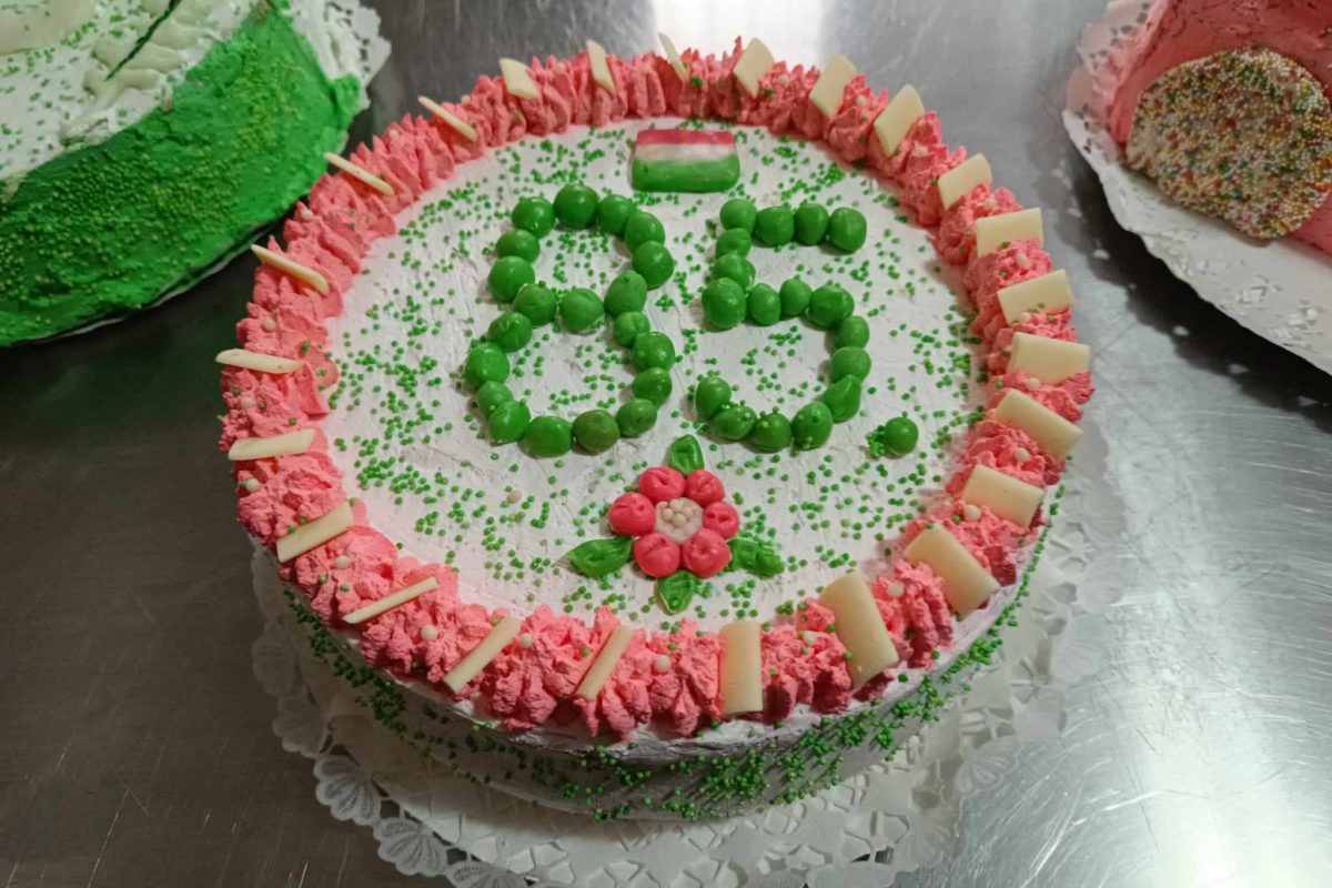 Kerek születésnapi torta zöld és rózsaszín színekkel díszítve. A tetején zöld színben a „85” szám látható, amelyet rózsaszín cukormáz vesz körül fehér csokoládédarabokkal. A szám alatt egy kis virágminta látható, a torta pedig egy csipke szalvétán, egy fémfelület tetején.