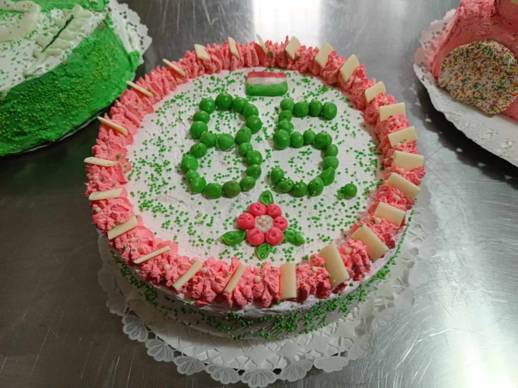 Születésnapi torta, zöld cukormázzal "85" számmal, rózsaszín cukormáz szegéllyel és fehér csokoládé darabokkal körülvéve. A tortát zöld szórással és rózsaszín virágmintával díszítették, és fémes felületre szalvétára helyezték.