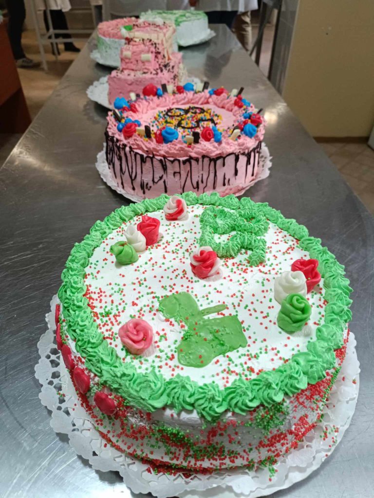 Négy színes, díszített torta sora egy rozsdamentes acél asztalon. Az előtérben lévő torta zöld és piros cukormázzal díszített, rózsamintákkal, míg a többiek a háttérben különböző élénk mintákat és feltéteket mutatnak be.