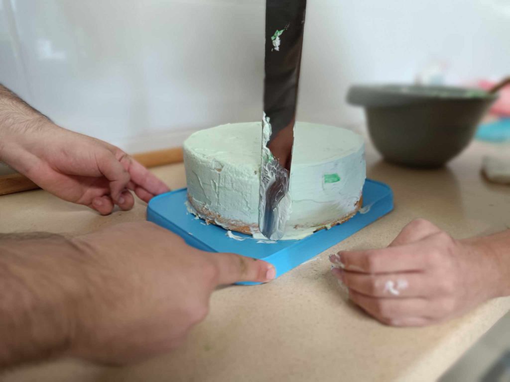 A bal oldalon két kéz látható, amelyek egy tortát tartanak egy kék tálcán. Egy másik kéz a jobb oldalon van, cukormázzal. Egy nagy kés van a közepén, vágja vagy simítja a zöldre fagyott tortát. Egy keverőtál van a háttérben egy pulton.
