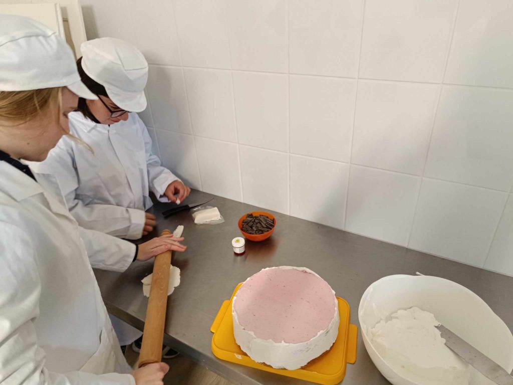 Két fehér köpenyes és kalapos ember dolgozik egy konyhában. Az egyik sodrófával kinyújtja a fehér tésztát; a másik apró darabokat vág. Egy nagy, kerek, rózsaszín és fehér torta áll a közelben, egy tál csokoládédarabokkal és kisebb edényekkel egy rozsdamentes acél pulton.