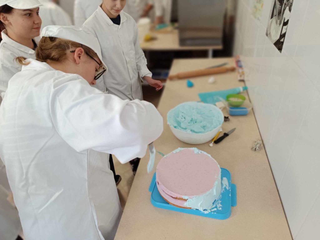Egy fehér laborköpenyt és sapkát viselő személy világoskék cukormázzal habosít egy tortát. A torta kék tálcán pihen egy pulton. Két másik ember is hasonló öltözékben nézi. Különféle sütőeszközök és több cukormázzal ellátott tál található a pulton.