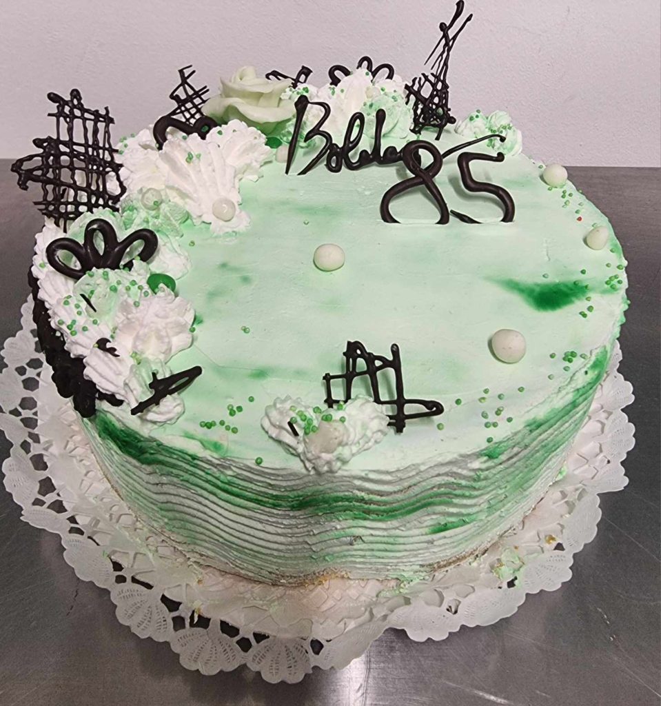 Kerek torta fehér és zöld cukormázzal, csokoládé díszítéssel és fehér virágokkal díszítve. A tortán „Bobka 85” felirat olvasható csokoládé betűkkel. Rozsdamentes acél felületen, csipkés fehér szalvétán ül.